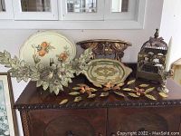Photo showing all decorative items on top of a wooden cabinet including plaques, floral garlands, decorative birdcage, and wall shelf.