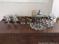 Wide view of all glass barware items including stemmed glasses, tumblers with gold trim, a glass plate, ribbed glass stirrer and metal shaker on wooden surface