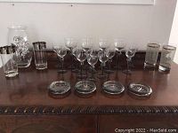 Set of various glassware including glasses with silver trim, black stemmed glasses, clear glasses, and four glass coasters on wooden surface.