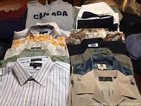 Top view photo showing a grey Canada hoodie, multiple folded shirts with collars and patterns including stripes, camouflage, and solid colors.