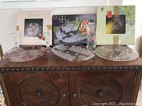 Three glass platters on wooden cabinet, two boxes in background with floral platter images.