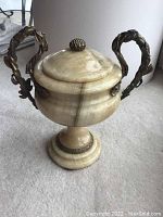 Front angled view of the marble vase showing its cream veining and two bronze leaf motif handles with lid and finial detail.