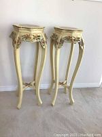 Pair of vintage cream-colored wooden plant/display stands with carved floral details and curved legs on carpeted floor against wall.