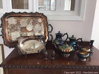 Full view of all silver plated items including ornate large tray, smaller tray on stand, tea service, and salt and pepper shakers.