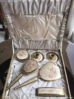 Overview of vintage vanity set displayed inside open box lined with satin fabric, showing embroidered floral tops and gold-tone accents on all items.