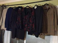 Photo showing several women's jackets and blouses hanging on a rod, colors include beige, brown, black, and navy with some floral or patterned designs.