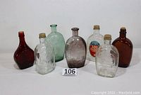 Photo of seven vintage glass syrup bottles in various colors including amber, clear, green, purple, and brown. Bottles are approximately 8.5 inches tall with embossed and labeled features.