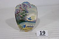 Full view of hand painted ceramic wall plaque depicting two flying blue birds by the lake with blooming trees and gold detailing on top edge.