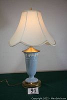 Front view of blue Lenox porcelain lamp with off-white scalloped fabric shade on green surface, showing base and floral decoration near top.
