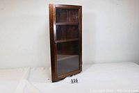 View of wooden corner display cabinet with closed glass door and four shelves inside.