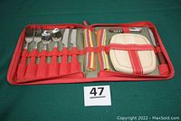 Opened red fabric trifold case showing stainless steel flatware with red handles, including forks, spoons, and knives, two striped cloth napkins, small wooden cutting board, and corkscrew bottle opener inside.