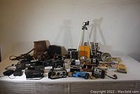 Wide image showing the entire lot including multiple vintage cameras, cases, flash units, tripod, and camera manuals scattered on white surface