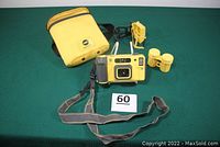 Full lot view showing the camera, yellow carrying case with strap, sports finder accessory, dual film holder, and camera neck strap on a green surface with a lot number card.