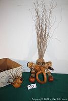 Two whimsical folk art faces made of natural plant materials including dried gourds, seed pods, nuts, and curly twigs. Larger face stands taller with long twig hair and prominent facial features; smaller face is compact with twig hair and round facial elements.