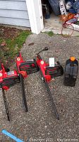 Three red Homelite electric chainsaws with attached power cords and a container of bar oil, arranged on concrete ground near a garage.