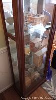 Front and side view of tall display cabinet showing wood frame, multiple glass shelves, and some boxed contents inside.