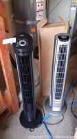 Two tower fans side by side, black Seville Classic and silver Lasko, with box in background