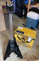 Full view of black Dean Flying V guitar on stand next to Fender Mustang 1 box
