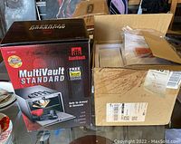Photo of GunVault MultiVault Standard safe box and cardboard box with time recorder and accessories on glass table.