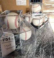 Image showing 6 signed baseballs in individual clear plastic cases stacked on a surface with a maxsold tag stating 'Signed Baseballs'.