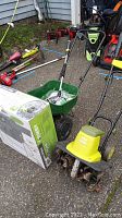 Wide view of yard care equipment including electric tiller Sun Joe, green spreader cart, boxed Chapin sprayer, and various garden tools and hose fittings laid on pavement.
