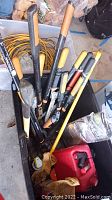 Various Fiskars hand tools with wooden handles in a bin including pruners and loppers, yellow crowbar, leather gloves, red gas can visible.