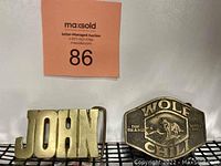 Photo showing both brass belt buckles side by side: one inscribed 'JOHN' and another embossed with 'WOLF CHILI' and a wolf design.