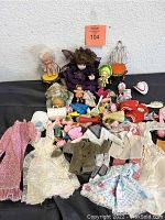 Wide view of assorted dolls including porcelain and plastic dolls with various doll clothing and accessories spread out on table.