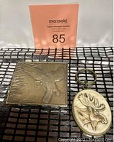 Photo showing the flying bird belt buckle and Canadian moose keychain together on a grid surface.