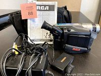 RCA vintage video camera with attached hand strap, showing lens, viewfinder, and various controls. Includes power adapter, AV cable, two rechargeable batteries, and instruction manuals.