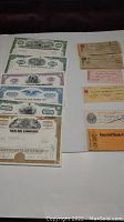 Photo shows 7 stock certificates and multiple cheques arranged on a table.
