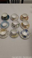 Nine assorted English bone china tea cups and matching saucers arranged on a table, displaying a variety of floral and decorative patterns.