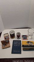 Full view of lot showing mini travel scale, two tin can coin banks, Aurora lighter, Bernard Shaw book, cigarette pack, and glass.