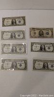 Photo showing all seven silver certificates laid out on a flat surface, six $1 bills and one $5 bill.