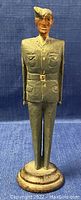 Front view of one hand carved wooden figurine of a Royal Canadian Trenton Cadet in green uniform standing on a small pedestal base.