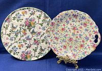 Two porcelain cake plates photographed side by side on dark blue cloth. Left plate is Barker Bros Tudor Ware with birds and flowers; right plate is Chintz with floral pattern and handles.