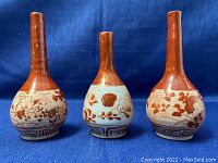 Three Asian hand painted porcelain bud vases with red floral and geometric designs shown from the front on a blue background.