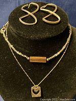Photo showing pair of infinity-shaped earrings, beaded choker with engraved clasp, and chain with two pendants on black display.