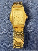 Front view of gold-tone rectangular case, white dial, and expansion band on blue background.