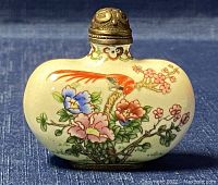 Front view of oval opium snuff bottle hand painted with floral and bird designs on cream enamel surface with metal cap.