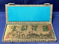 Rectangular Chinese metal plaque with calligraphy displayed inside an open silk box with blue patterned lining and turquoise cushion interior.
