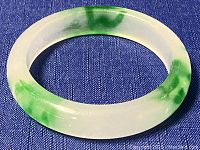 Jade bangle bracelet with green veining and a translucent whitish base on a blue fabric background, side view showing curved surface.