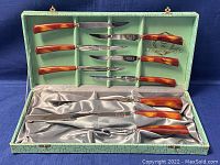 Full carving set in open green presentation case showing 9 pieces including 3 large knives, carving fork and 4 smaller knives with butterscotch Bakelite handles.