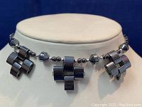 Close-up front view of the necklace showing three cross-shaped hematite stone beads with smaller round and faceted beads on a white display stand.