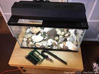 Glass aquarium tank with black lid and light, filled with seashells and sea rocks on wooden table with filter parts beside it.