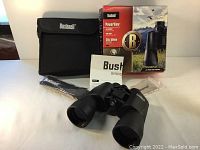 Bushnell binoculars displayed with original packaging, carrying case, neck strap, and lens cloth on a white surface.