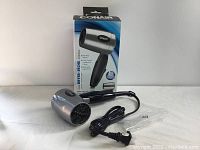 Full view of Conair 124AC silver travel hair dryer with foldable handle and black power cord, alongside original retail box.