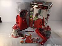 NutriSlicer base with crank handle attached, three stainless steel drums, food holder, and original packaging box arranged on tabletop.