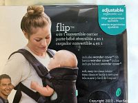 Photo of product front packaging showing woman carrying infant in black ergonomic baby carrier.
