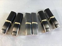 Photo showing six L'Oréal Paris Gold Addiction Colour Riche lipsticks in black to gold gradient packaging with gold band, all sealed and arranged flat.